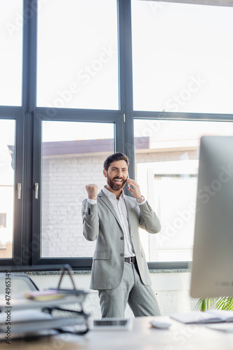 Excited businessman in suit showing yes gesture while talking on smartphone near computer in office.