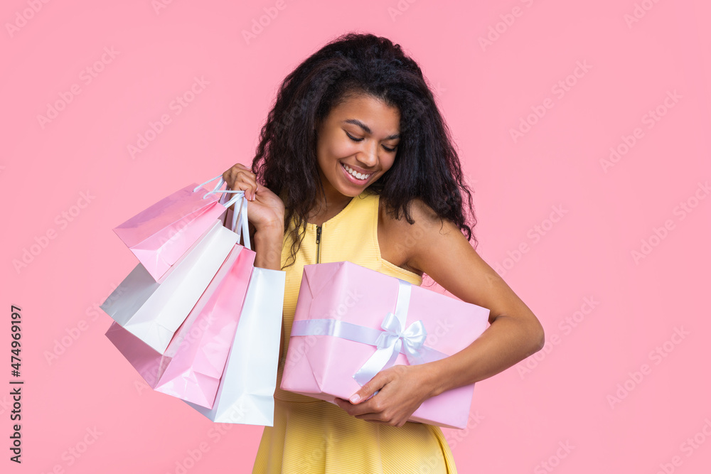 Portrait of beautiful girl with charming smile holding decorated gift box and a bunch of shoppers in hands, isolated over pastel pink background