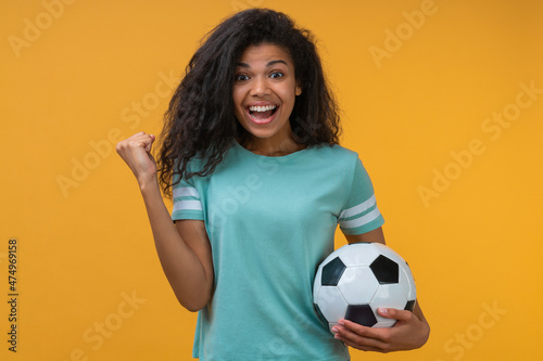 Portrait of euphoric happy football fan girl celebrating victory after betting at bookmaker's website, making winner's gesture clenching her fist while holding ball in hand