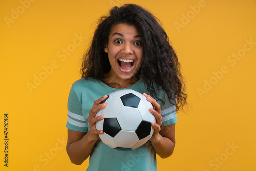 Studio shot of euphoric soccer fan girl cheering for her favourite team while holding the ball in hands, isolated over yellow background © wpadington