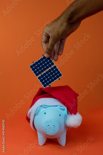 Hand putting photovoltaic solar panel in piggy bank with christmas santa claus hat isolated on orange background. Concept image.