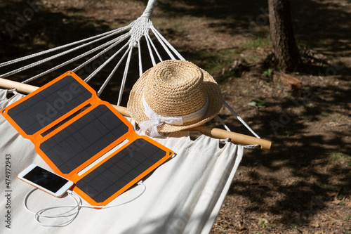 Smartphone is charging from solar battery in summer camp. Clean energy for using in camping. Gadgets and sun hat are lying in a hammock.