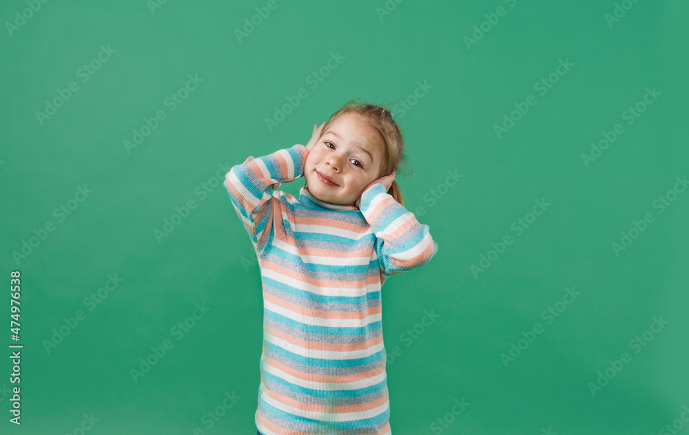 a little beautiful girl in a striped jacket covers her ears with her hands on an isolated green background a little beautiful girl in a striped jacket covers her ears with her hands on an isolated green background