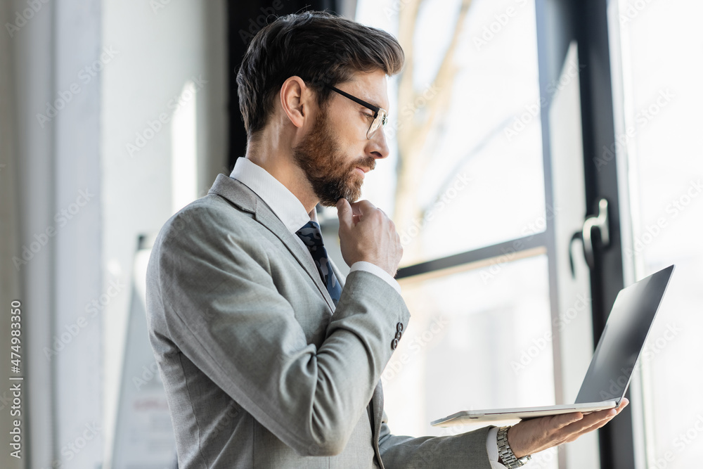 Side view of businessman in suit and eyeglasses looking at laptop in office. Side view of businessman in suit and eyeglasses looking at laptop in office.