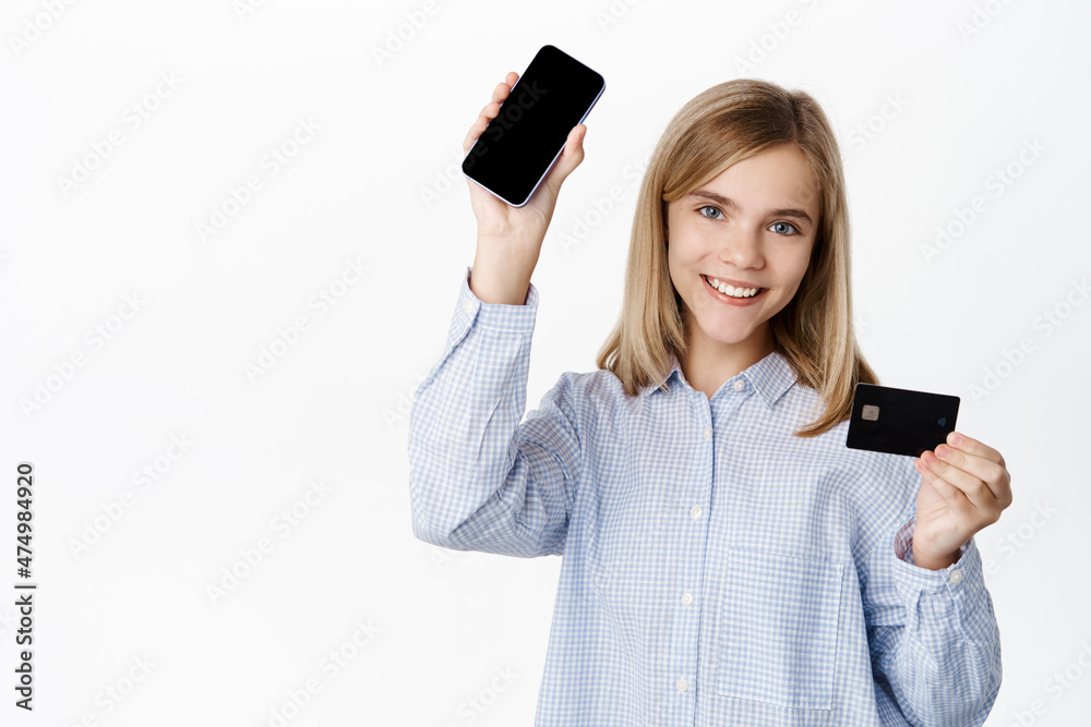 Cute little girl, small blond child showing credit card and smartphone app interface, mobile screen, standing over white background Cute little girl, small blond child showing credit card and smartphone app interface, mobile screen, standing over white background