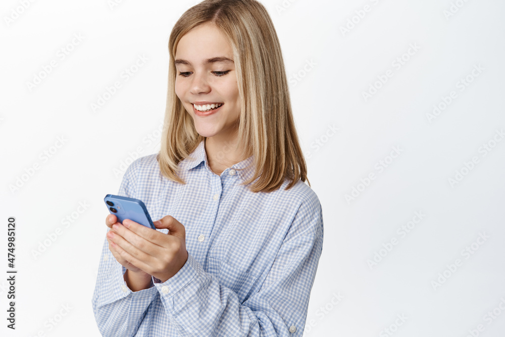 Cute blond girl, teen using mobile phone, chatting or scrolling social media app, standing over white background Cute blond girl, teen using mobile phone, chatting or scrolling social media app, standing over white background