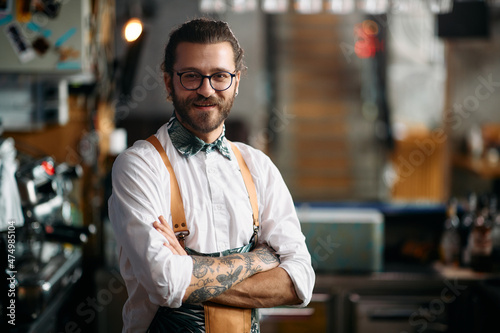 Portrait of happy barista stands with arms crossed at pub and looks at camera.