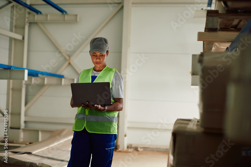 African American female warehouse worker uses laptop at storage compartment.