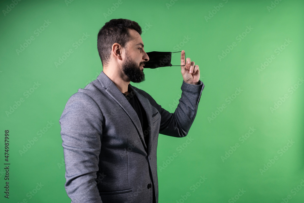 Elegant man of mixed arabic features wearing suit on green background as businessman takes off his face mask symbolizing the end of the pandemic Elegant man of mixed arabic features wearing suit on green background as businessman takes off his face mask symbolizing the end of the pandemic