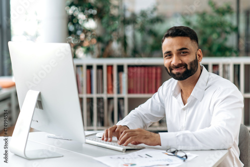 Friendly smart confident businessman of Indian ethnic, manager or ceo with beard sitting in office, wearing white shirt, using computer, texting with clients, taking notes, looking at camera, smiling