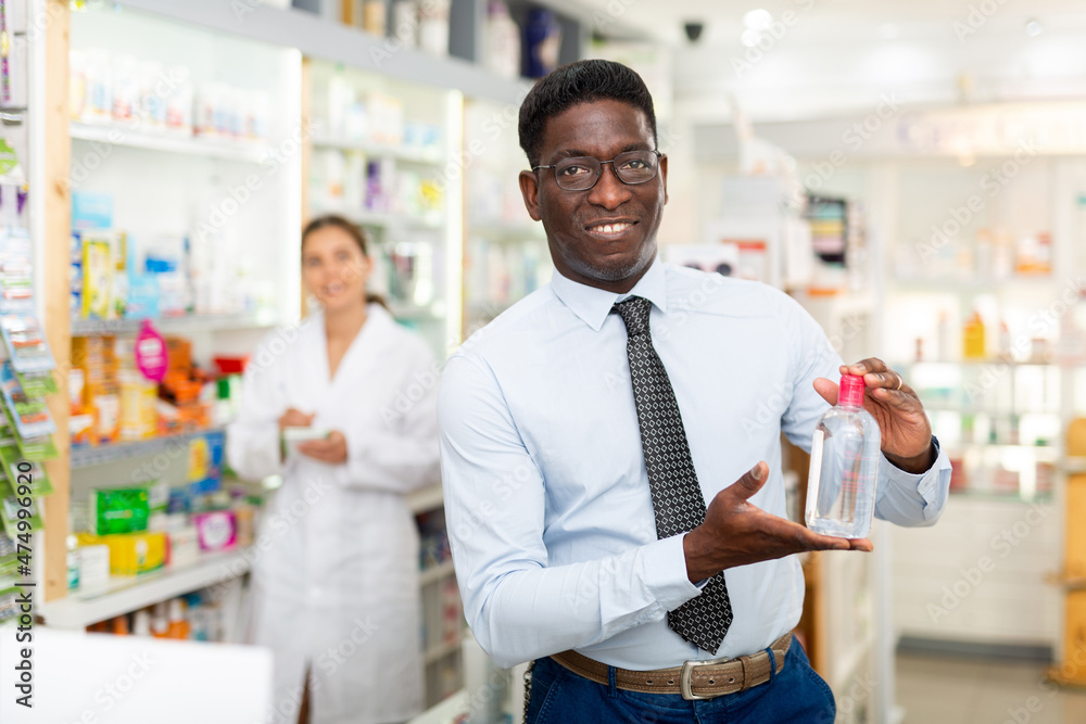 African-american man standing in salseroom of drugstore and holding bottle of cosmetic product in hand. Pharmacist working in background.