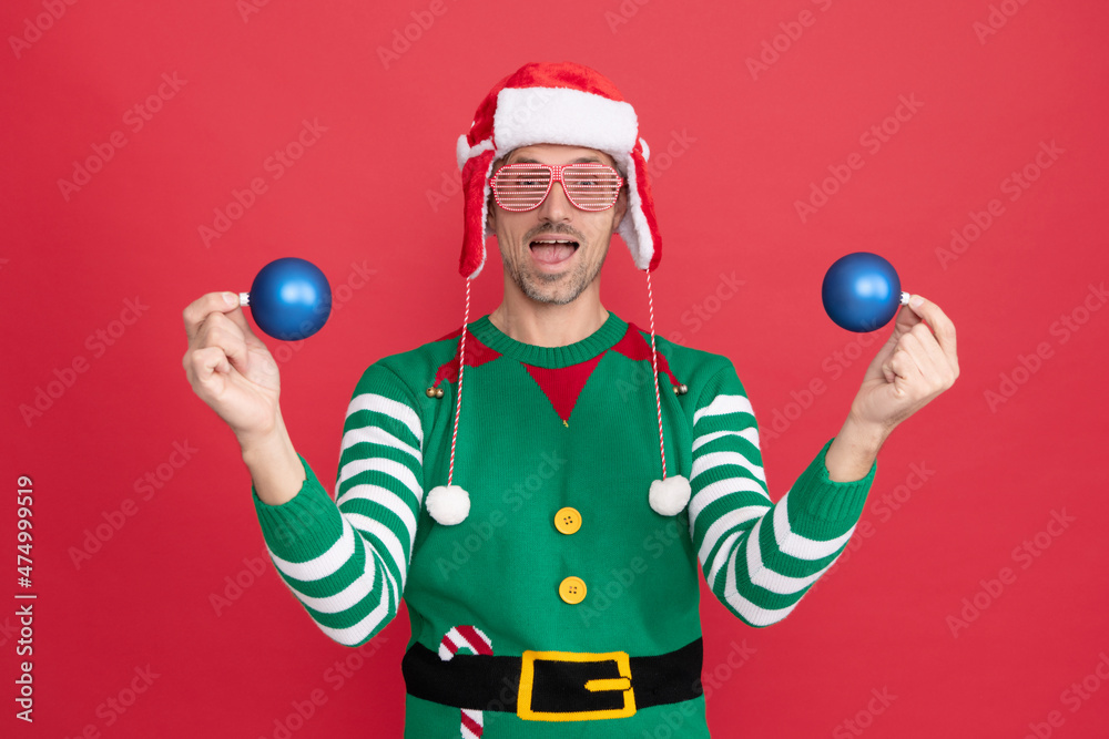 amazed man in elf costume and santa claus hat. guy in party glasses hold xmas decoration balls