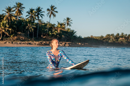 Photography Portrait of blond surfer girl on white surf board in blue ocean pictured from th