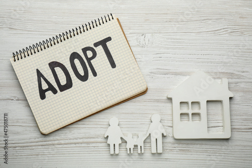 Family figure, house and notebook with word Adopt on white wooden table, flat lay photo