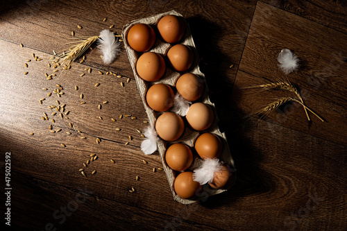 A dozen farm fresh organic brown eggs with scattered feathers and wheat against a textured background with side light and top down view.
