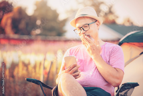 Gray beard mature Man sit in the flowers farm outdoors and chitchat on online dating app in mobile phone. Happy retirement man in Pink Shirt and eyeglasses, Hat at Sunset. valentine's day concept photo