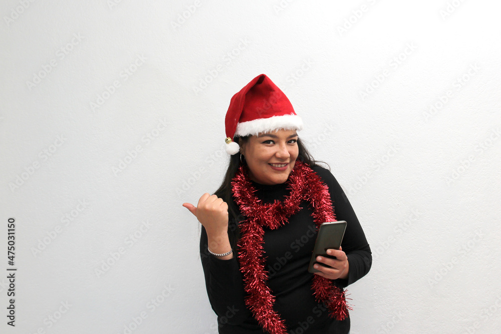 Latin adult woman with Christmas hat and red garland as scarf shows her cell phone that she uses for contests, promotions, calling her family with a video call happy and excited to celebrate Christmas Latin adult woman with Christmas hat and red garland as scarf shows her cell phone that she uses for contests, promotions, calling her family with a video call happy and excited to celebrate Christmas