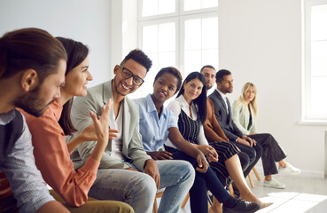 Diverse group of young people sitting in row in modern office and talking. Happy multi-racial men and women having interesting discussion during business meeting or psychological training