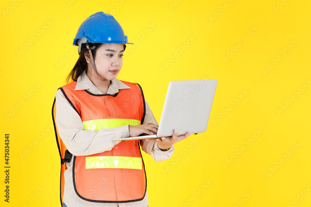 Studio shot of Asian female professional engineering foreman manager wear hard helmet and reflective safety vest stand smiling holding laptop computer working on industrial site on yellow background Studio shot of Asian female professional engineering foreman manager wear hard helmet and reflective safety vest stand smiling holding laptop computer working on industrial site on yellow background