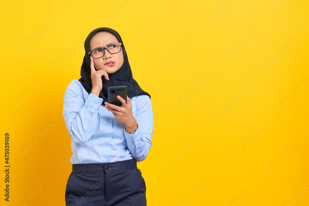 Portrait of pensive young Asian woman using a mobile phone and looking up isolated on yellow background Portrait of pensive young Asian woman using a mobile phone and looking up isolated on yellow background