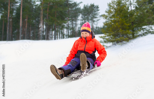 childhood, sledging and season concept - happy little girl sliding down on snow saucer sled outdoors in winter over snowy park or forest background