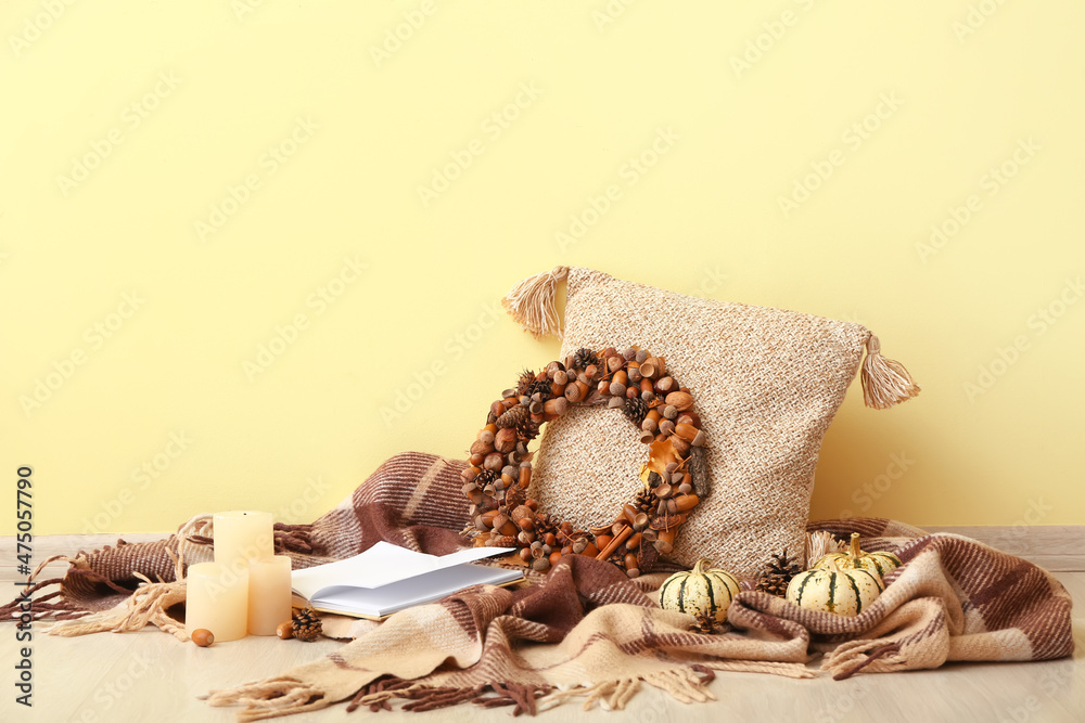 Fototapeta premium Beautiful acorn wreath, pillow, pumpkins, book and warm plaid near yellow wall