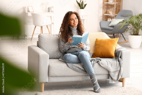 Young African-American woman with book sitting on sofa at home