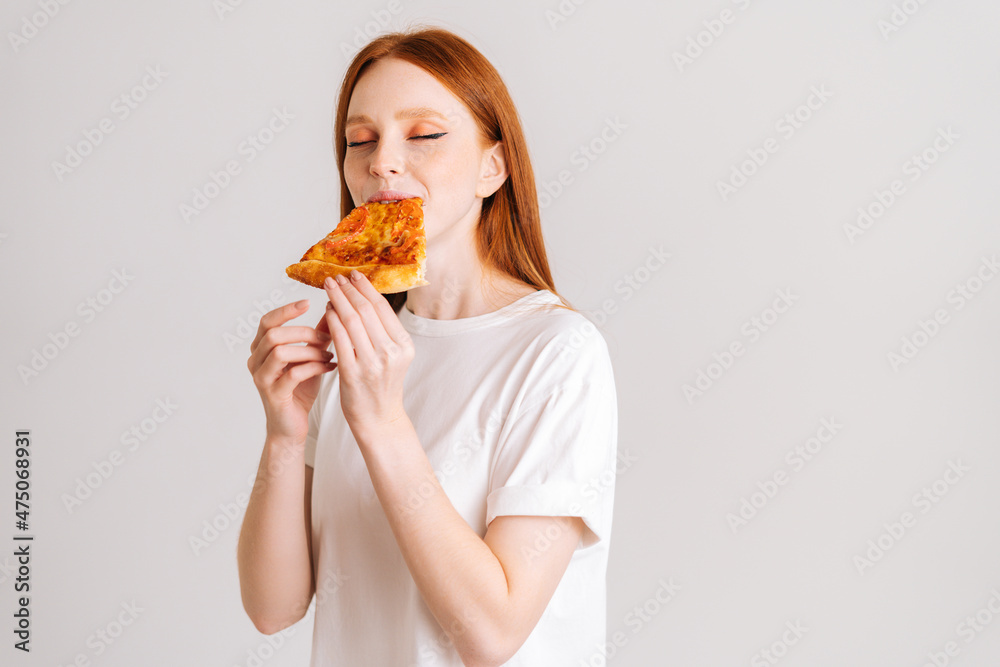 Studio portrait of cheerful young woman with closed eyes appetite eating delicious pizza standing on white isolated background. Pretty redhead female eating tasty meal. Studio portrait of cheerful young woman with closed eyes appetite eating delicious pizza standing on white isolated background. Pretty redhead female eating tasty meal.