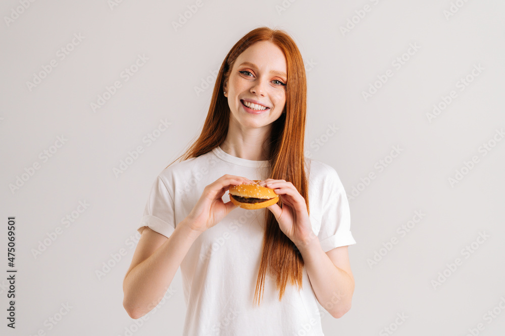 Studio portrait of smiling attractive redhead young woman holding tasty burger and looking at camera standing on white isolated background. Happy cute female eating delicious hamburger. Studio portrait of smiling attractive redhead young woman holding tasty burger and looking at camera standing on white isolated background. Happy cute female eating delicious hamburger.