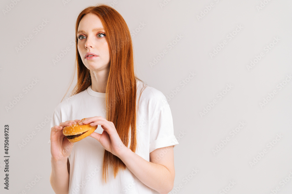 Studio portrait of shocked pretty young woman with disgust holding burger, looking away, standing on white isolated background. Confused cute redhead female eating hamburger.