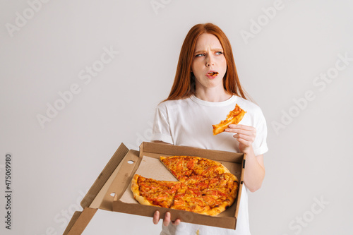 Studio portrait of dissatisfied young woman eating bad quality pizza holding box in hands looking away standing on white isolated background. Pretty redhead female eating unappetizing meal.
