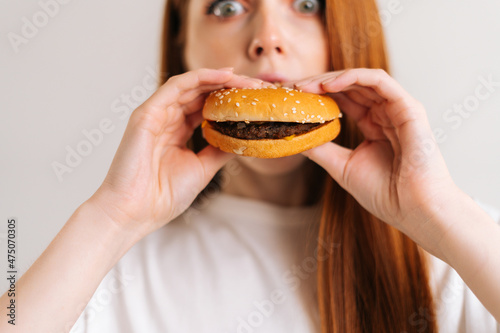 Close-up face of amazed pretty young woman with eyes wide open pleasing bite of appetizing delicious burger looking at camera, standing on white isolated background in studio, front view, portrait.