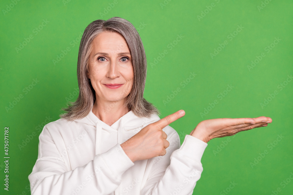 Photo of cute confident senior lady wear white hoodie pointing finger holding arm empty space smiling isolated green color background Photo of cute confident senior lady wear white hoodie pointing finger holding arm empty space smiling isolated green color background