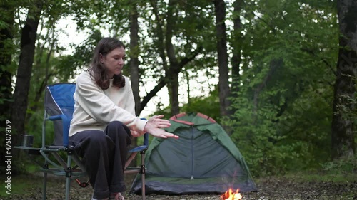A woman sits in a folding chair by a campfire in a camp. Tent on a hike. Wild life in nature