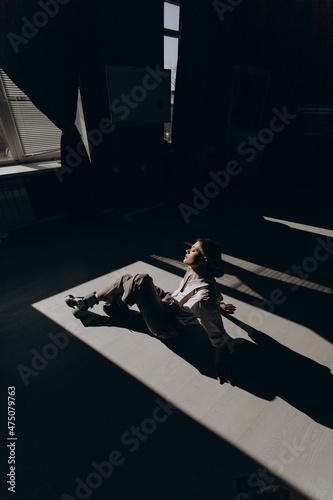Portrait of fashion retro girl in white shirt, suspenders and brown wide pants sitting on floor in the sun in room