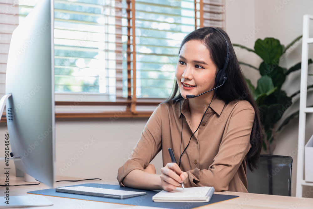 Asian beautiful woman wearing earphone and talking on microphone looking at laptop and writing, student woman learning online and write on notebook, stay at home Asian beautiful woman wearing earphone and talking on microphone looking at laptop and writing, student woman learning online and write on notebook, stay at home
