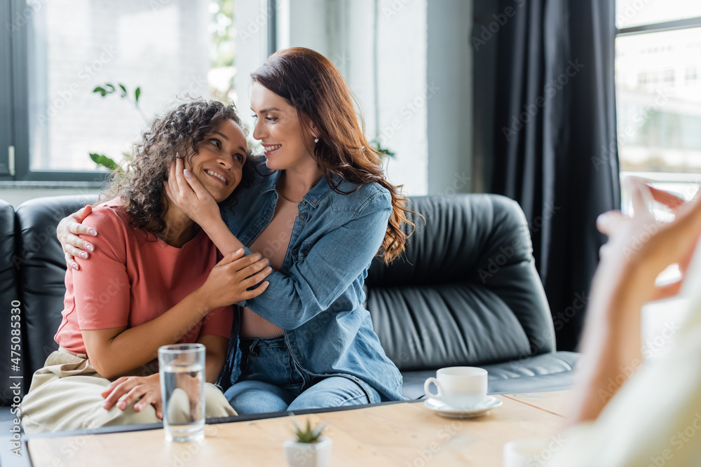 couple of interracial lesbian women hugging on couch near blurred psychologist in consulting room couple of interracial lesbian women hugging on couch near blurred psychologist in consulting room
