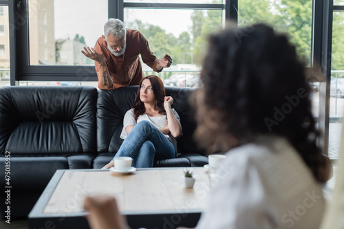 middle aged man gesturing while talking to offended wife during visit to psychologist on blurred foreground