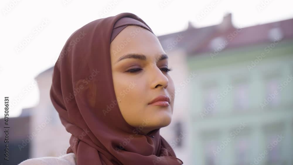 A young beautiful Muslim woman looks around in a street in an urban area - colorful buildings in the blurry background - face closeup A young beautiful Muslim woman looks around in a street in an urban area - colorful buildings in the blurry background - face closeup