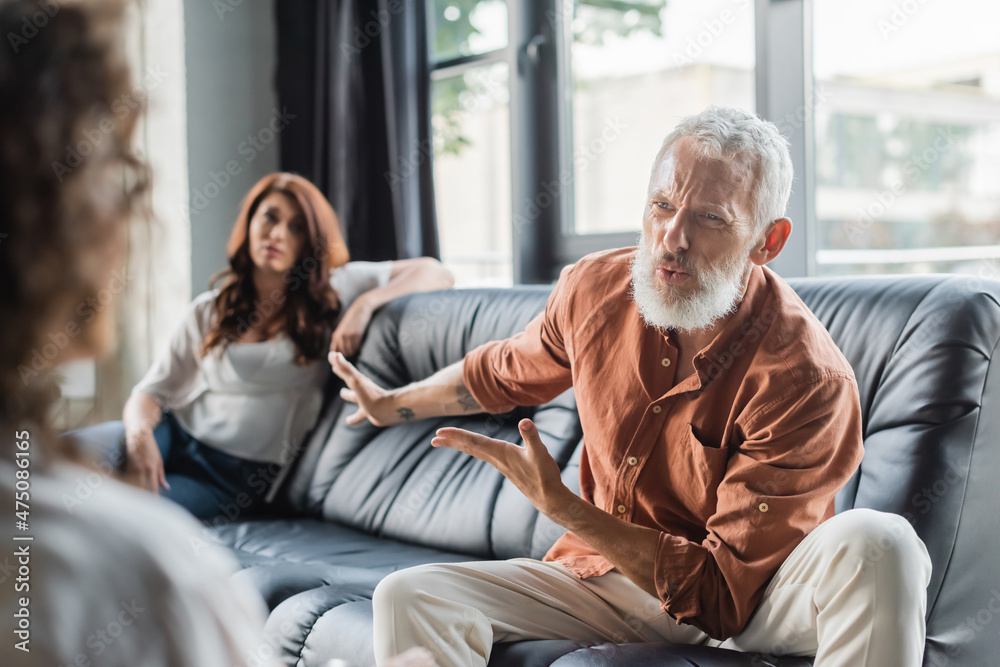 displeased man pointing at upset wife while talking to blurred african american psychologist displeased man pointing at upset wife while talking to blurred african american psychologist