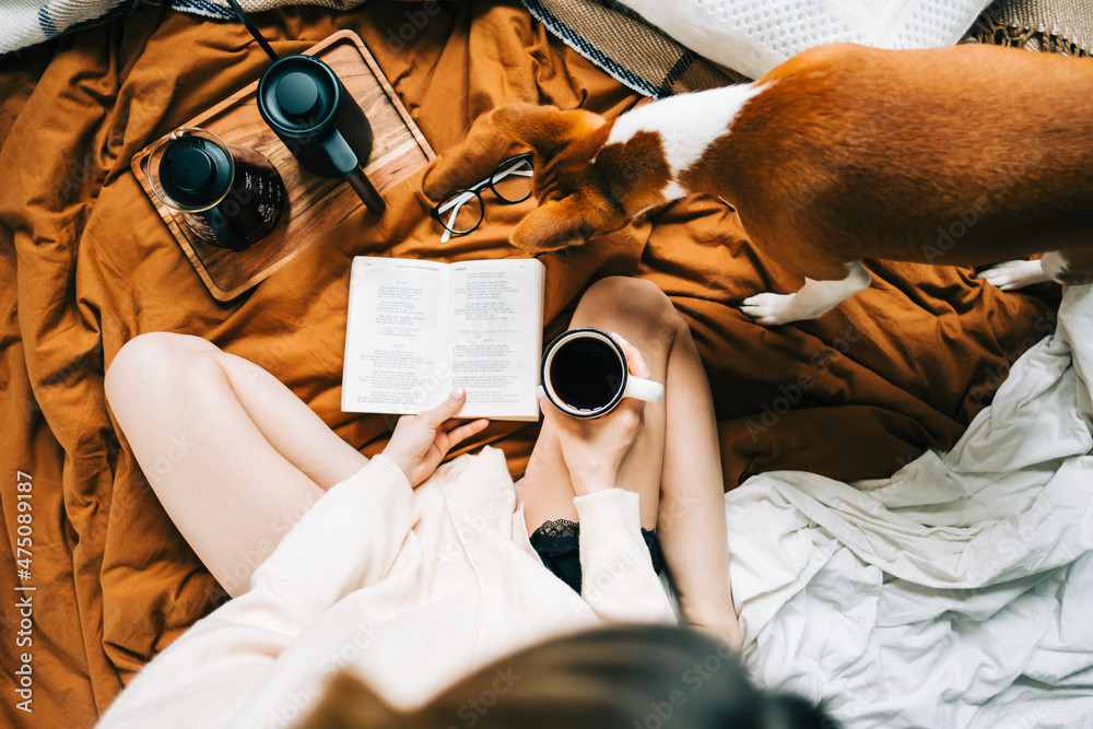 Caucasian woman drinking coffee in the morning and reading book on the bed at home with her pet, coziness and comfort concept.