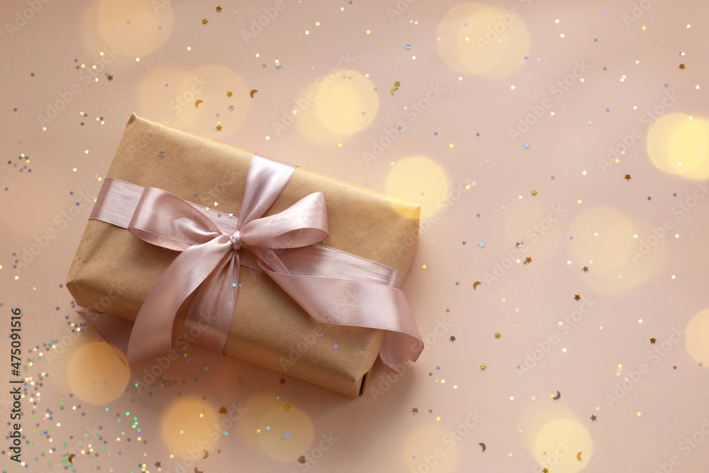 Fototapeta premium Birthday or New Year's eve present in kraft paper with pink ribbon on a soft beige background with glitter and confetti. Xmas composition. Flat lay. Happy holidays celebration and giving love concept