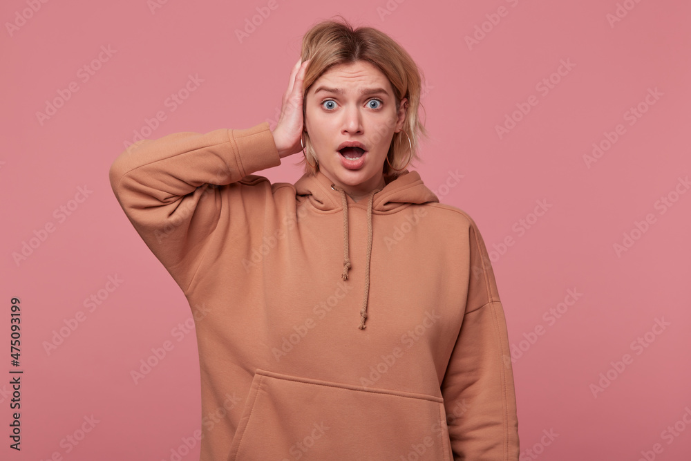 Studio portrait of young blonde woman with curly hair, wears beige hoodie looks into camera with shocked facial expression, keeps hand on head and mouth widely opened, isolated over pink background Studio portrait of young blonde woman with curly hair, wears beige hoodie looks into camera with shocked facial expression, keeps hand on head and mouth widely opened, isolated over pink background
