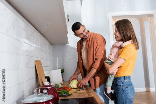 Positive woman hugging son while husband cooking salad in kitchen