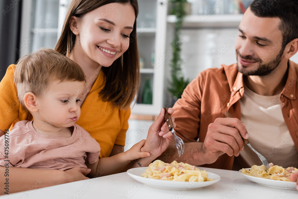 Smiling woman holding fork near pasta and family at home
