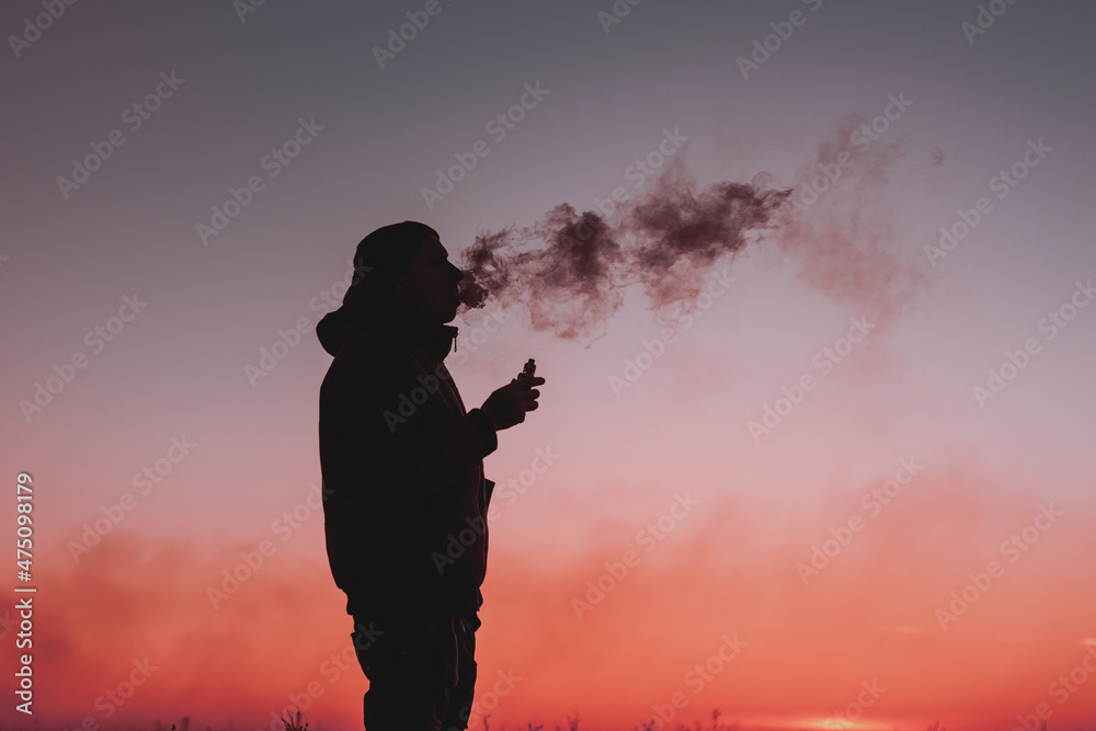 A man in a jacket smokes an electronic cigarette outdoors. Vaping on a sunset background. Alternative to analog cigarettes