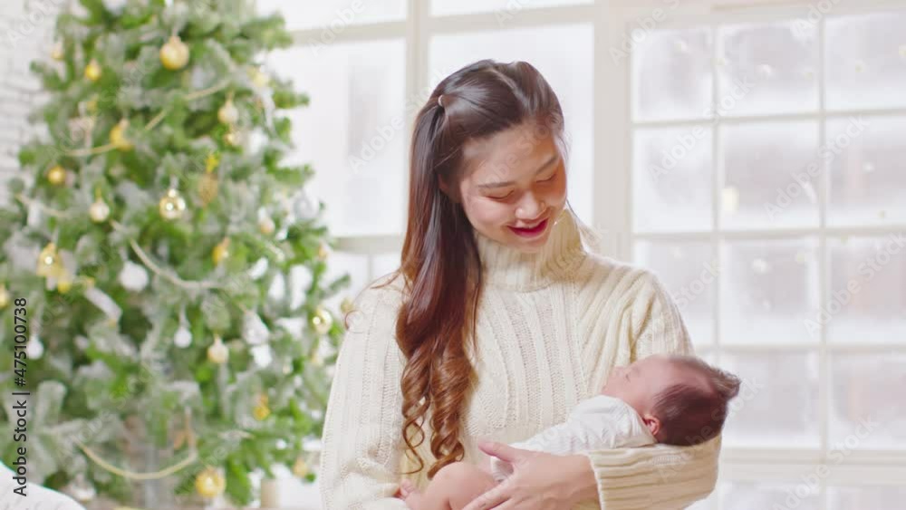 Beautiful Asian woman holding newborn baby in arms sit on bed at white cozy home with christmas tree in background.Happy infant baby sleep in mother arms safety and comfortable.Mom and Baby Concept Beautiful Asian woman holding newborn baby in arms sit on bed at white cozy home with christmas tree in background.Happy infant baby sleep in mother arms safety and comfortable.Mom and Baby Concept