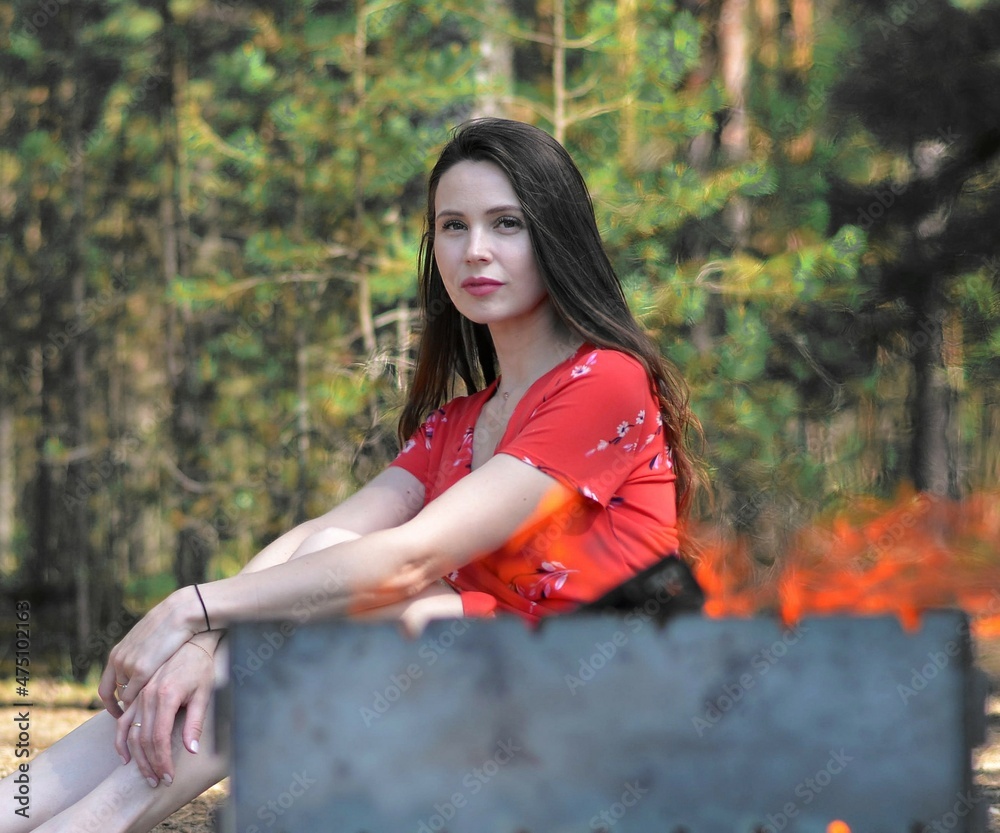 a young beautiful Caucasian woman is sitting at an iron barbecue on a picnic, looking at the viewer, long dark hair, in a red dress, clasped her knees, summer a young beautiful Caucasian woman is sitting at an iron barbecue on a picnic, looking at the viewer, long dark hair, in a red dress, clasped her knees, summer