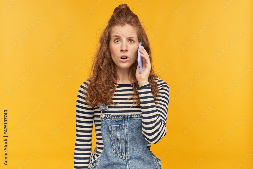 indoor studio shot of young ginger female with long red curly hair talking on the phone with confused, guilty facial expression, isolated over yellow background indoor studio shot of young ginger female with long red curly hair talking on the phone with confused, guilty facial expression, isolated over yellow background