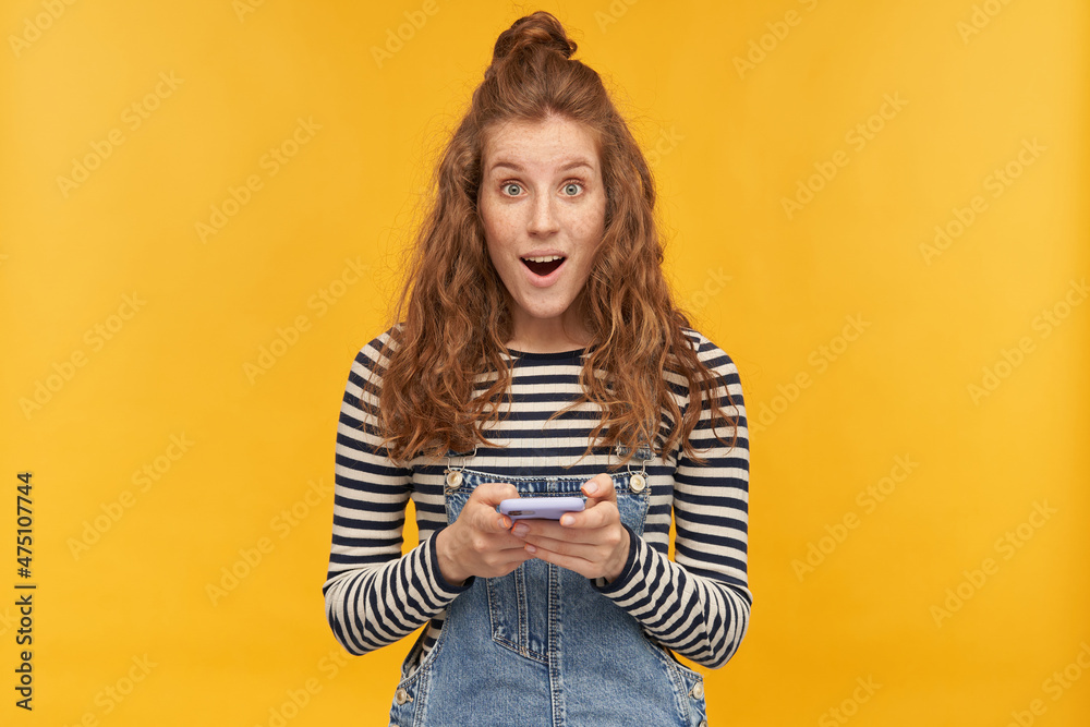 indoor studio shot of young ginger female with long red curly hair holding her phone looks directly into camera with shocked, surprised facial expression isolated over yellow background indoor studio shot of young ginger female with long red curly hair holding her phone looks directly into camera with shocked, surprised facial expression isolated over yellow background